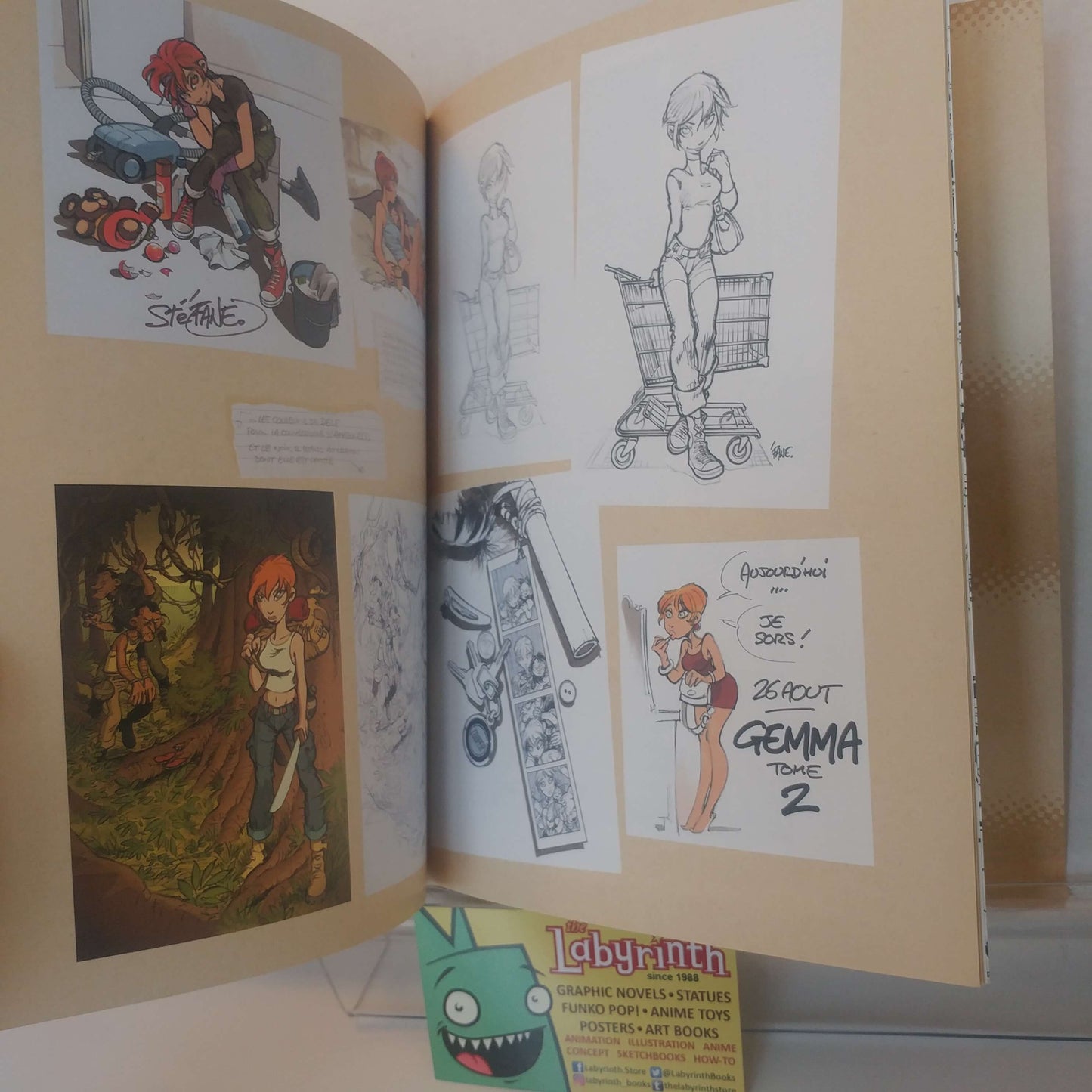 An open art book or sketchbook showing multiple illustrated panels, including character drawings and a comic-style panel, photographed on a tabletop with a small Labyrinth toy visible in front.