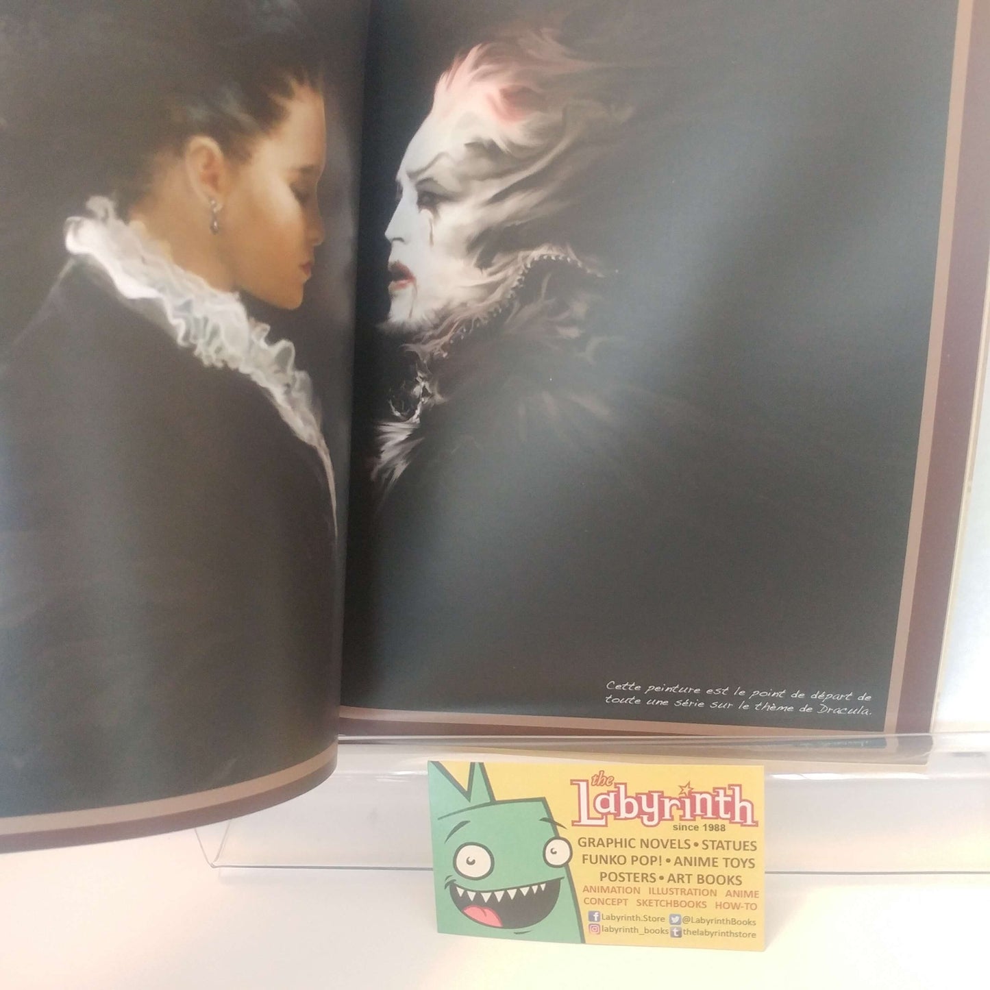 An open book or magazine spread showing a dark illustrated portrait of a woman on the left page and a white-faced figure on the right page. A small Labyrinth-themed card or box sits in front on a clear display stand.
