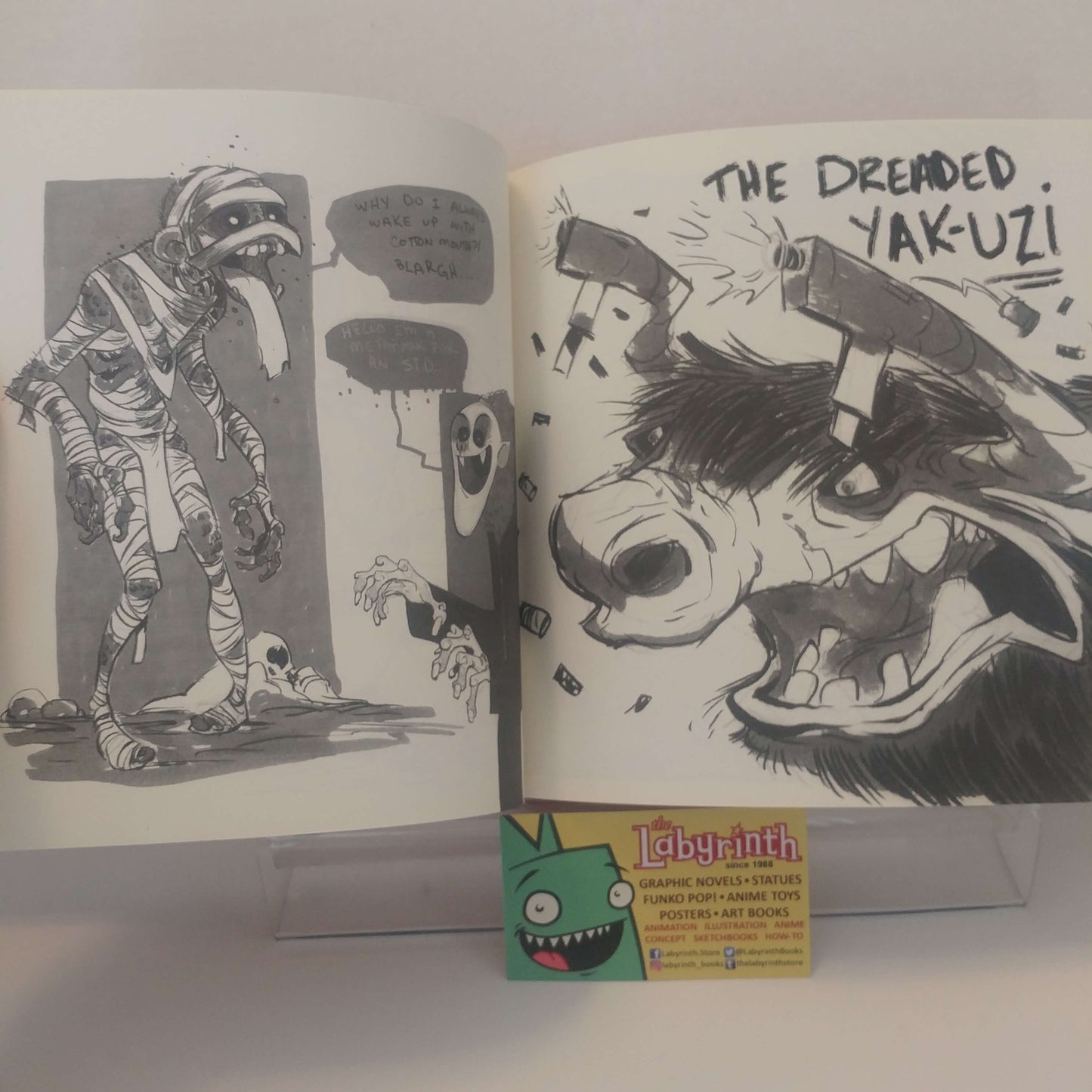 An open illustrated art book or comic spread on a white surface, showing black-and-white comic panels and a bold black ink illustration of a dog with the text 'THE DREMEID. YAK-UZ!'.