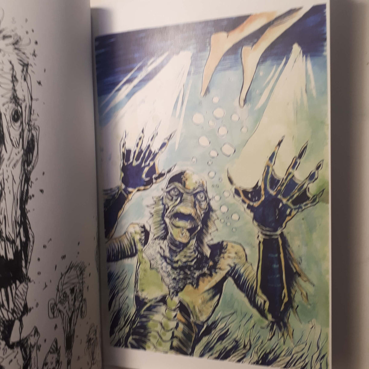 A close-up of an illustrated page showing a monstrous creature with clawed hands reaching upward in a blue-green underwater scene, with partial black line art visible on the left page.