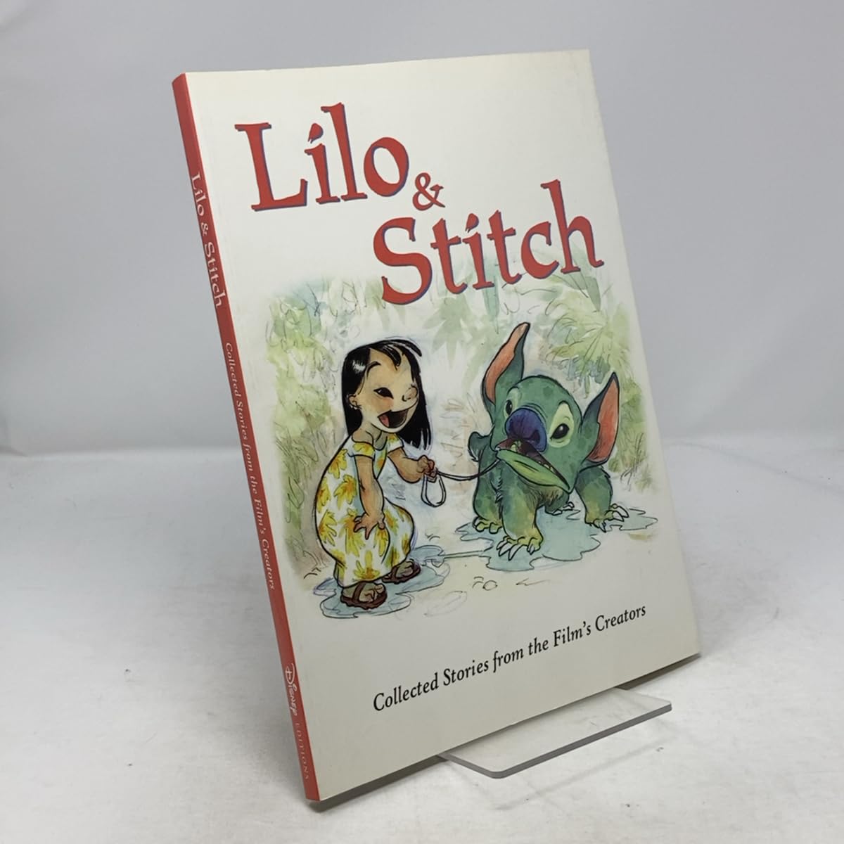 A hardcover book titled 'Lilo & Stitch' standing upright on a small clear display stand against a light gray background. The cover shows an illustration of Lilo and Stitch, with the title in large red letters and smaller text reading 'Collected Stories from the Film's Creators.