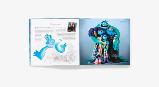 An open art book or magazine spread on a light gray background, featuring a blue creature illustration on the left page and a group photo of blue monster characters on the right page.