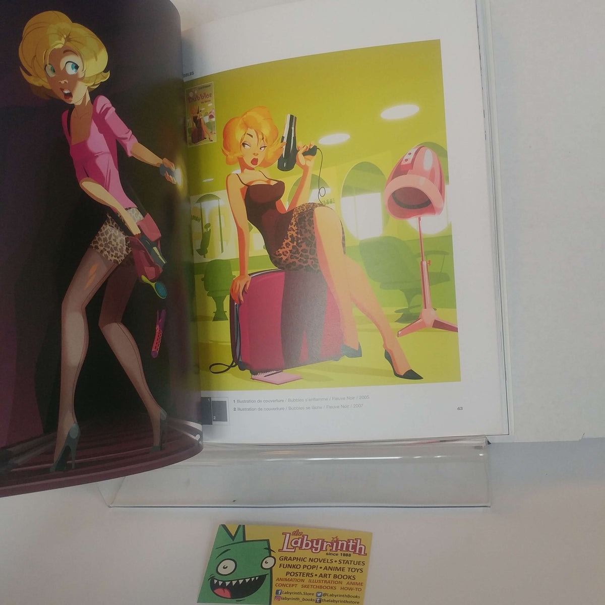 An open art book or magazine showing illustrated fashion imagery on the left page and a colorful fashion illustration on the right page, photographed on a white surface with a small card in front.