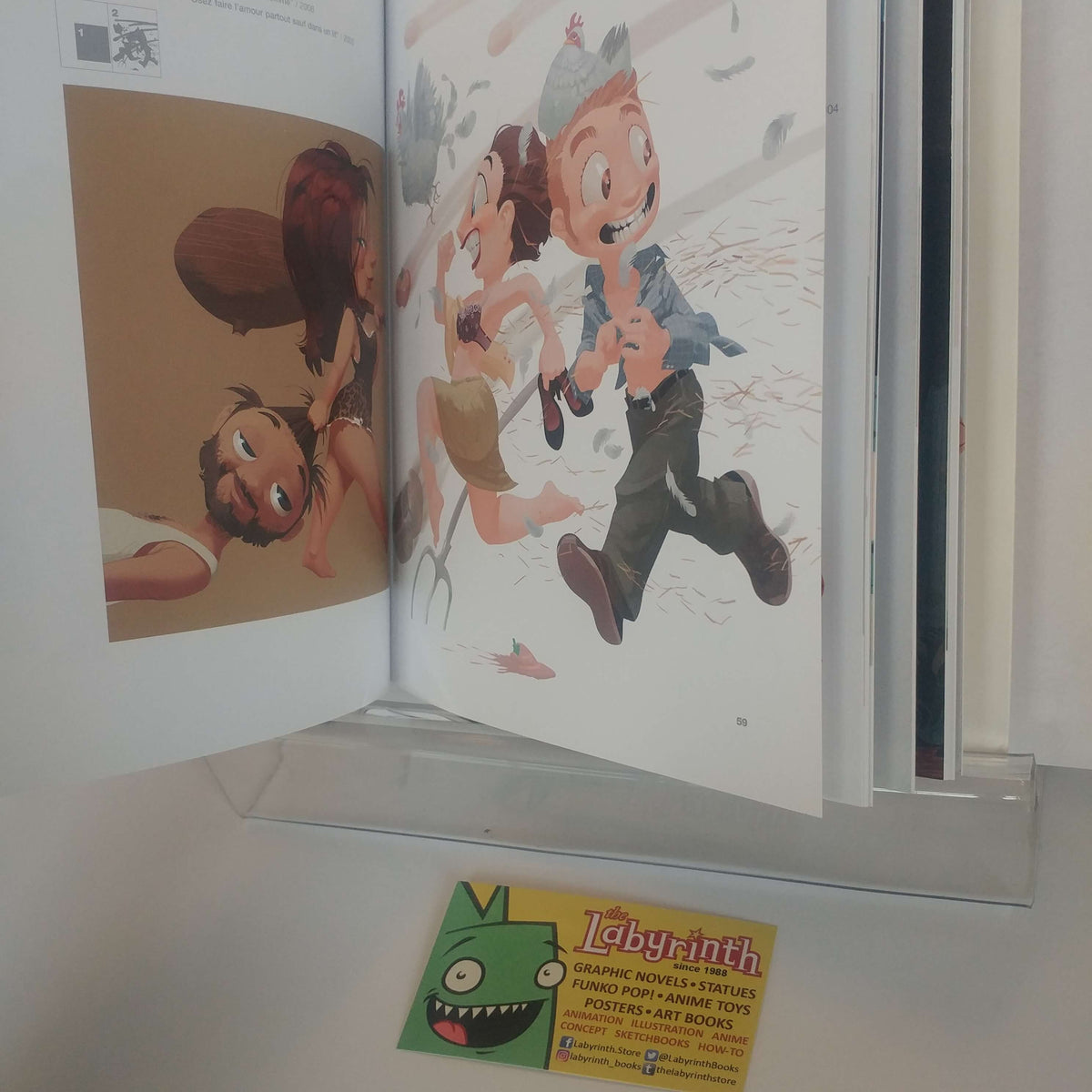 An open art book or comic volume displayed on a clear acrylic stand, showing illustrated pages with cartoon characters. A small card for 'Labyrinth' is visible in front of the book.