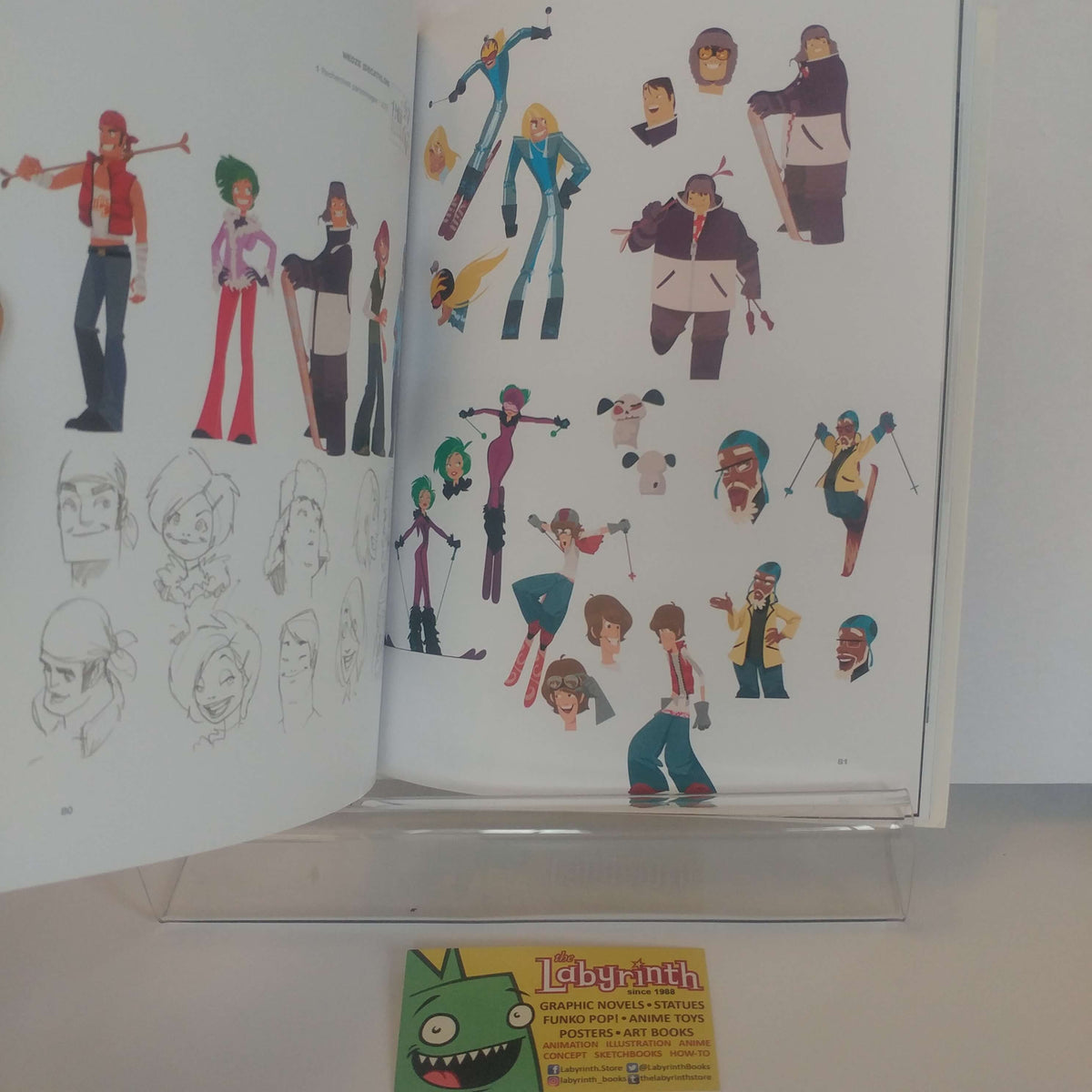 An open art book or character design portfolio displayed on a clear stand, showing multiple colorful character illustrations and sketch pages. A small yellow card with a green cartoon face and the word 'Labyrinth' is visible in front.