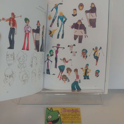 An open art book or character design portfolio displayed on a clear stand, showing multiple colorful character illustrations and sketch pages. A small yellow card with a green cartoon face and the word 'Labyrinth' is visible in front.