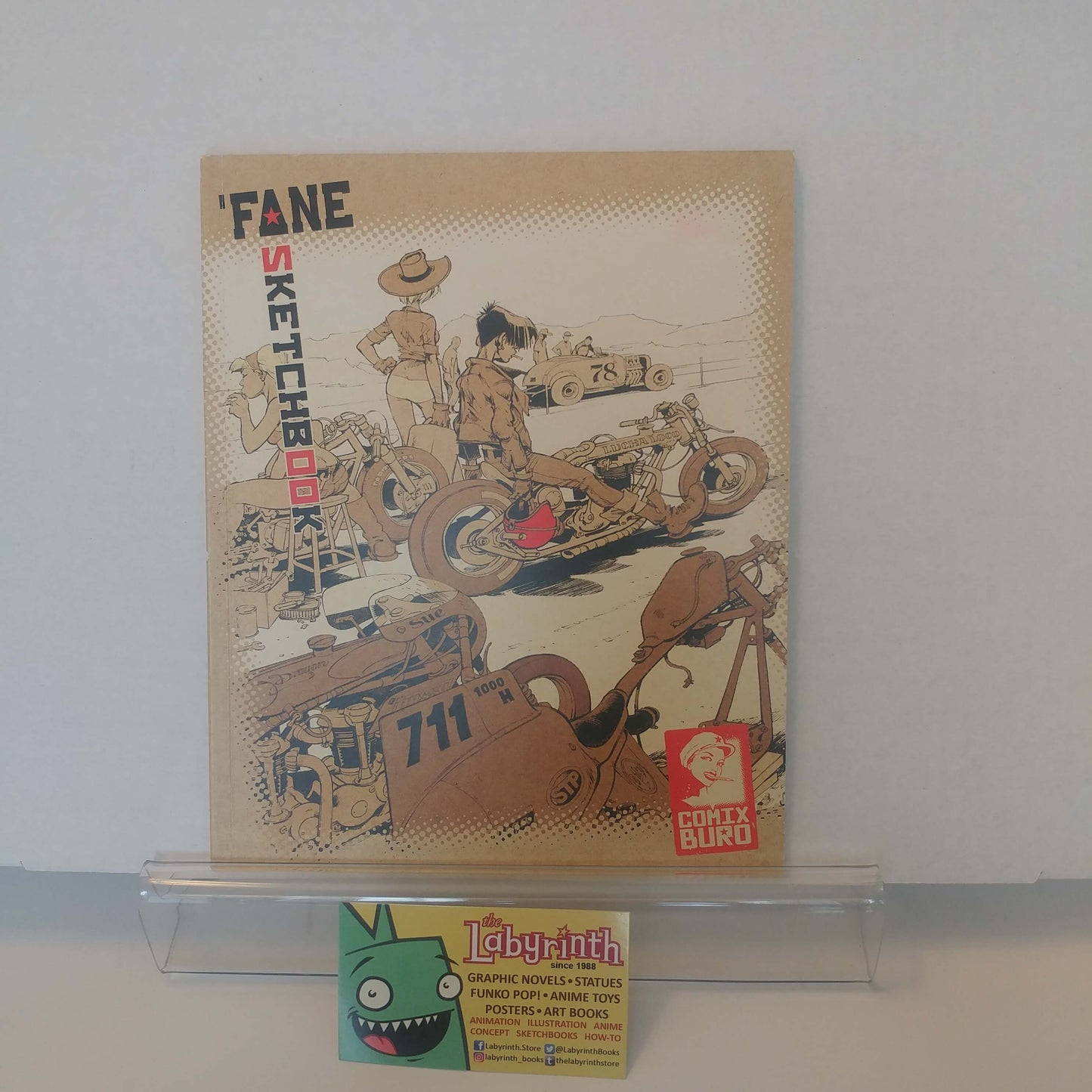 A vintage 'Fane Sketchbook' comic book by Conty Biro is displayed on a clear acrylic stand. The beige sketchbook features retro-style illustrations of a motorcycle, race car, and cowboy, with the title 'FANE SKETCHBOOK' printed on the front.