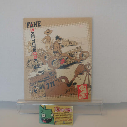 A vintage 'Fane Sketchbook' comic book by Conty Biro is displayed on a clear acrylic stand. The beige sketchbook features retro-style illustrations of a motorcycle, race car, and cowboy, with the title 'FANE SKETCHBOOK' printed on the front.