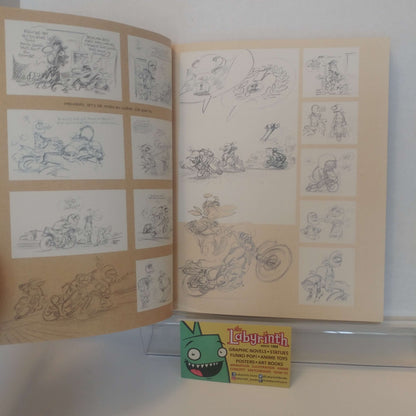 An open sketchbook displaying comic book character designs and storyboards, with a small 'Labyrinth' card placed in front on a clear display stand against a white wall.