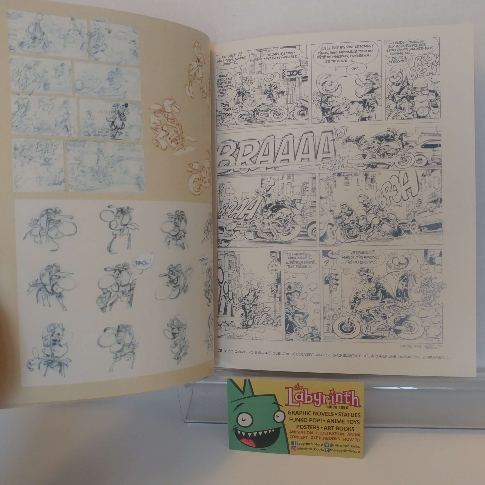 An open vintage comic book or art book showing two illustrated pages with black-and-white comic panels and blue sketch drawings on cream paper. A small Labyrinth card is placed in front on a clear stand against a white background.