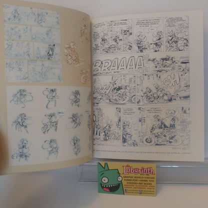 An open vintage comic book or art book showing two illustrated pages with black-and-white comic panels and blue sketch drawings on cream paper. A small Labyrinth card is placed in front on a clear stand against a white background.