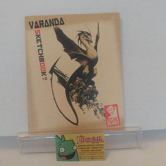 Varanda Sketchbook #2 from Conix Buro, displayed on a clear acrylic stand next to a Labyrinth trading card. The sketchbook features a beige background with a black illustration of a dragon and the title 'VARANDA SKETCHBOOK #2' printed on the cover.
