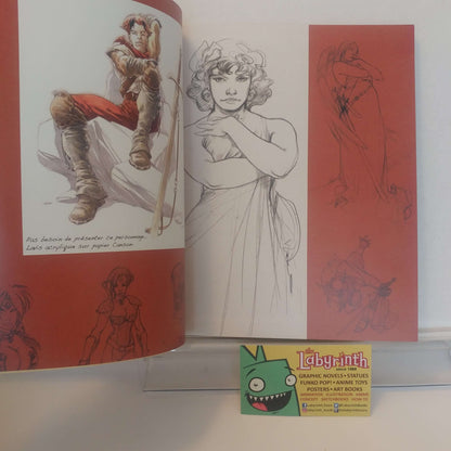 An open art book or sketchbook showing character illustrations and pencil sketches, with a small Labyrinth card placed in front on a white surface.