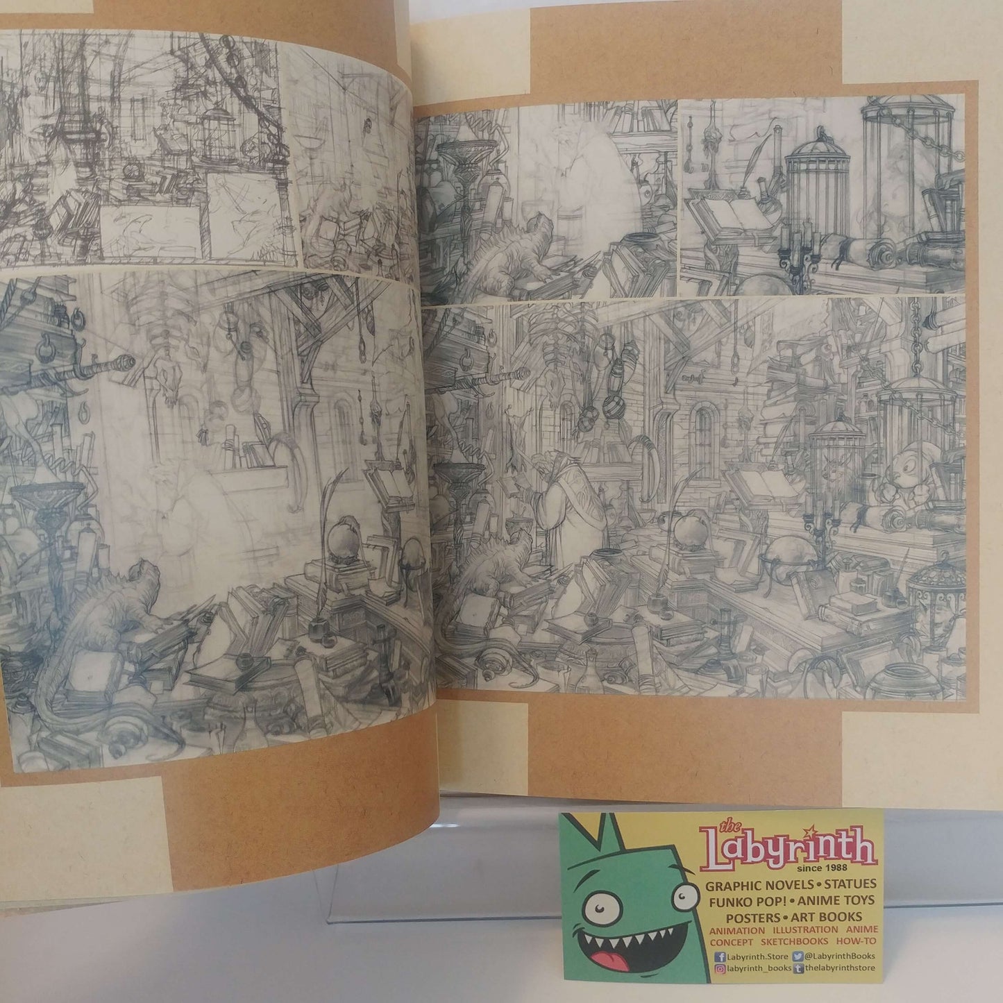 An open sketchbook or art book showing detailed pencil-style comic or animation scene drawings on cream paper. A small card at the bottom right reads 'Labyinth' and includes text about gaming, mystery figures, funding, anime toys, posters, and art books.