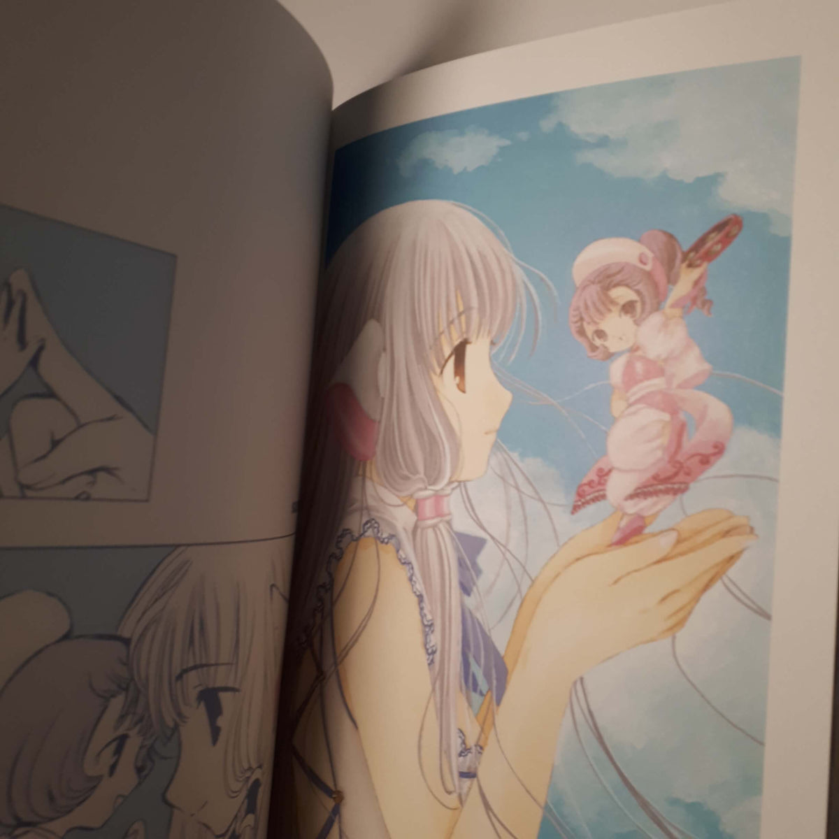 An open art book or manga volume showing a printed illustration on the right page of a pale-haired girl holding a small pink character against a blue sky background. from chobits your eyes only art book