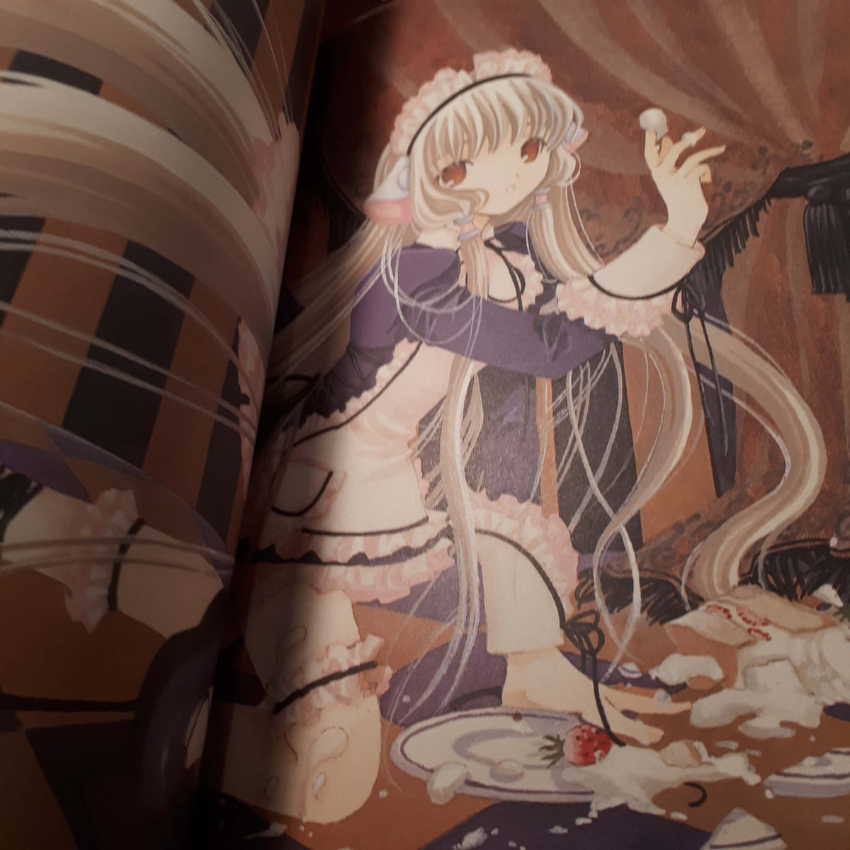 An open book or printed magazine showing an illustrated anime-style page with a long-haired female character in a purple and cream outfit, surrounded by warm brown tones and a plate with a strawberry. from chobits your eyes only artbook