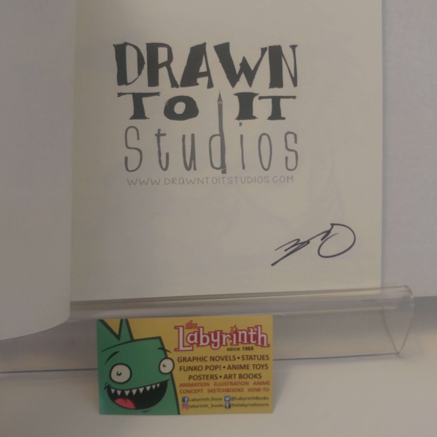 A signed copy of the 'Drawn to It Studios' comic book, featuring a black ink signature on the white cover. A small 'The Labyrinth' business card is resting on the clear acrylic stand in front of the book.