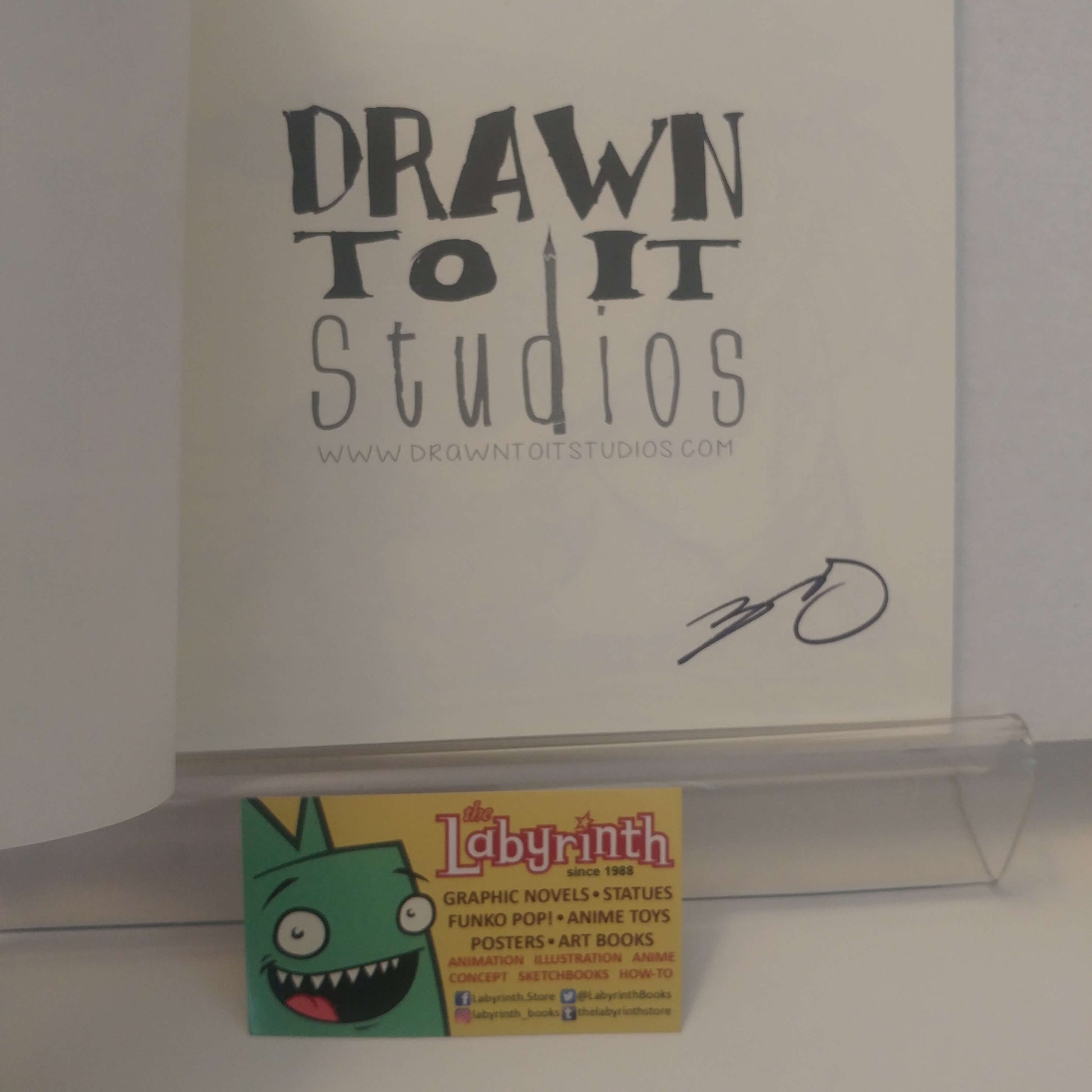A signed copy of the 'Drawn to It Studios' comic book, featuring a black ink signature on the white cover. A small 'The Labyrinth' business card is resting on the clear acrylic stand in front of the book.