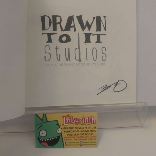 A signed copy of the 'Drawn to It Studios' comic book, featuring a black ink signature on the white cover. A small 'The Labyrinth' business card is resting on the clear acrylic stand in front of the book.