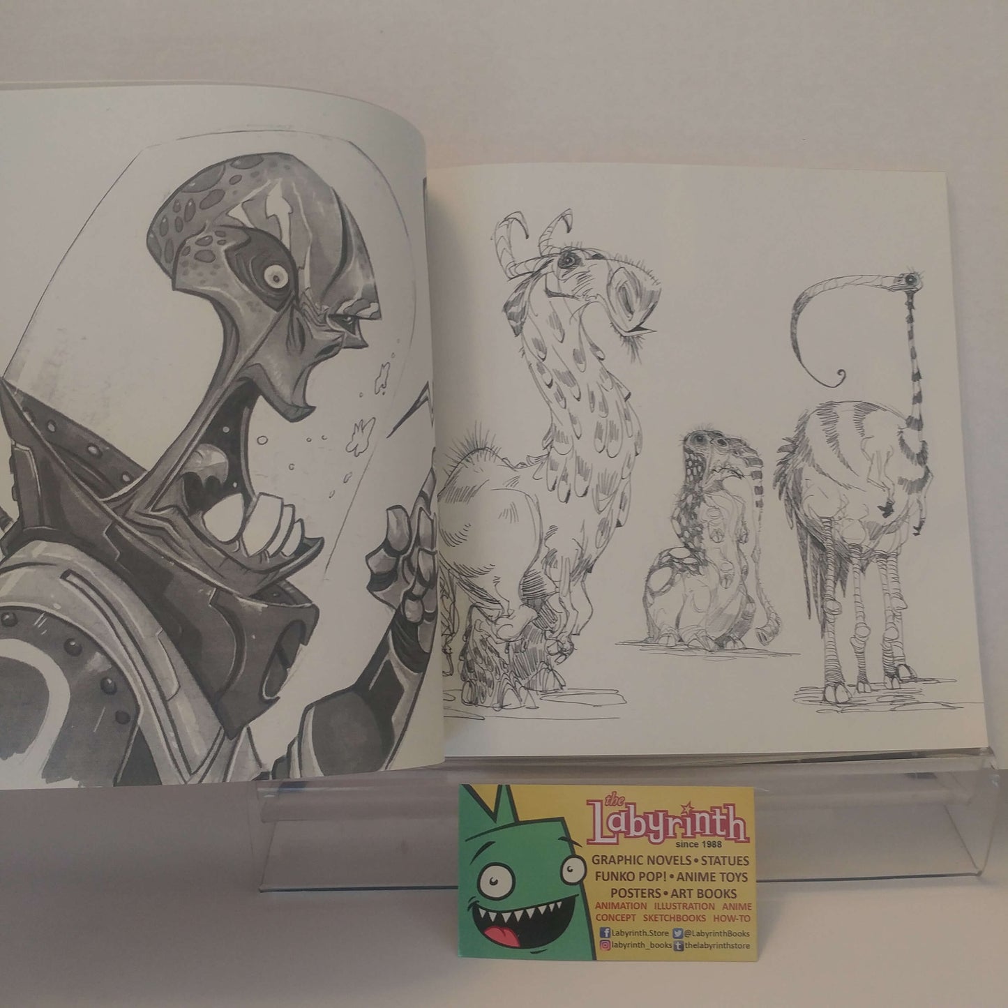 An open art book or sketchbook displayed on a clear acrylic stand, showing black-and-white character and animal illustrations. A small business card for Labyrinth is placed in front of the book.