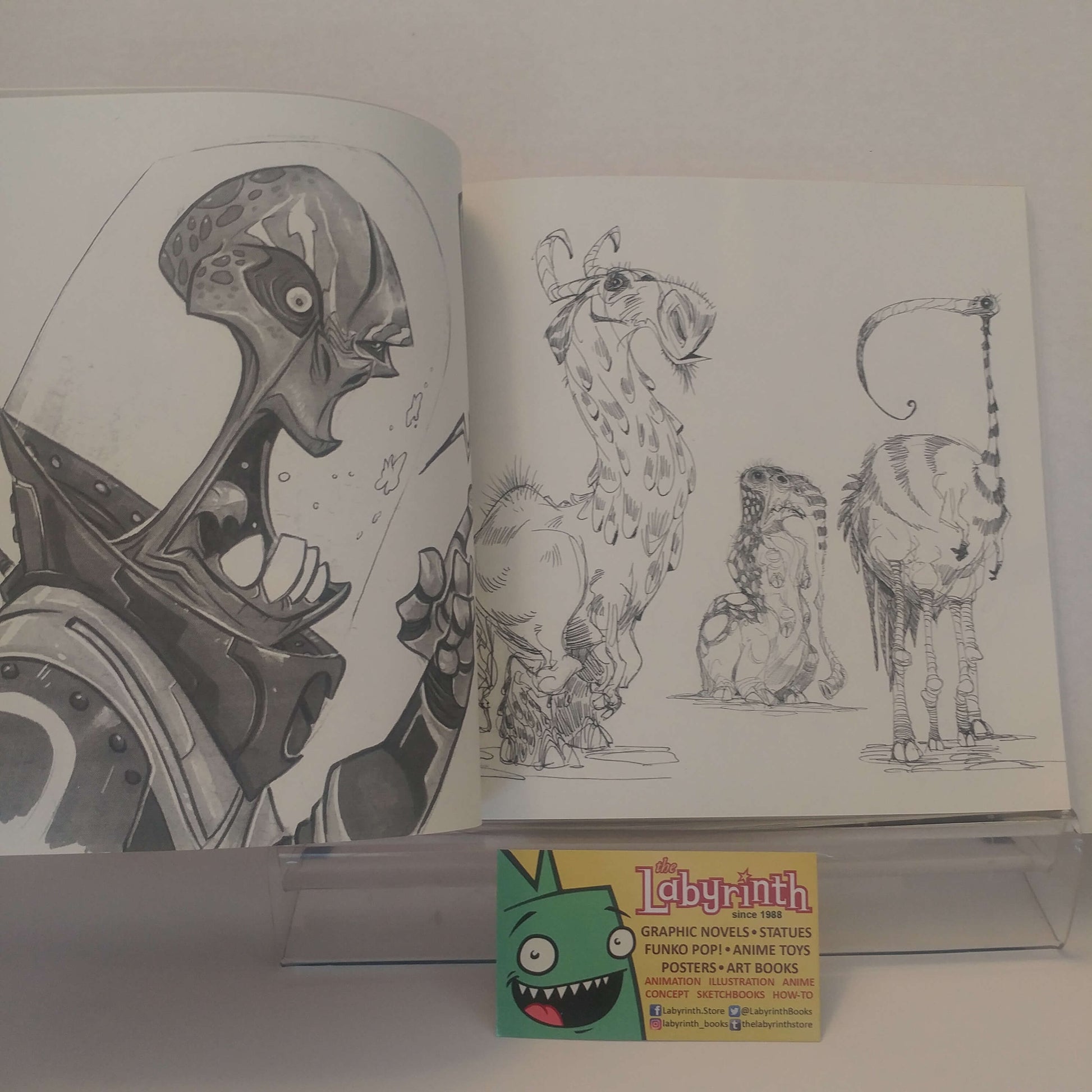 An open art book or sketchbook displayed on a clear acrylic stand, showing black-and-white character and animal illustrations. A small business card for Labyrinth is placed in front of the book.