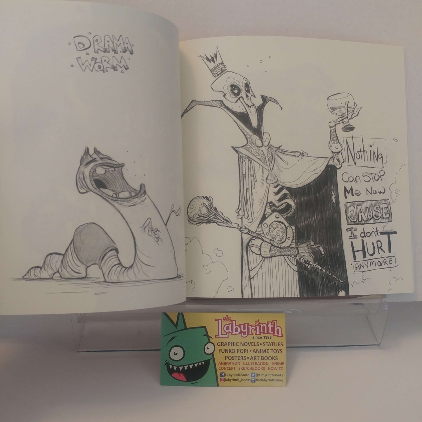 An open sketchbook or art print showing hand-drawn cartoon characters and handwritten text, displayed on a clear stand with a small Labyrinth card in front. The artwork appears to be the product being shown.