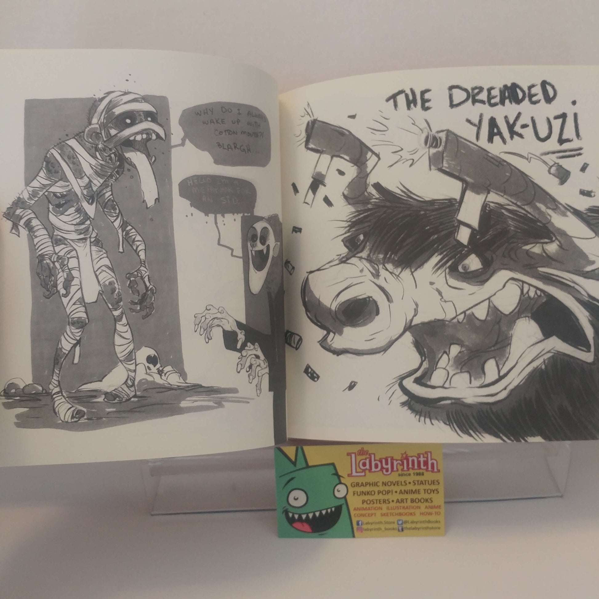 An open illustrated art book or comic spread on a white surface, showing black-and-white comic panels and a bold black ink illustration of a dog with the text 'THE DREMEID. YAK-UZ!'.