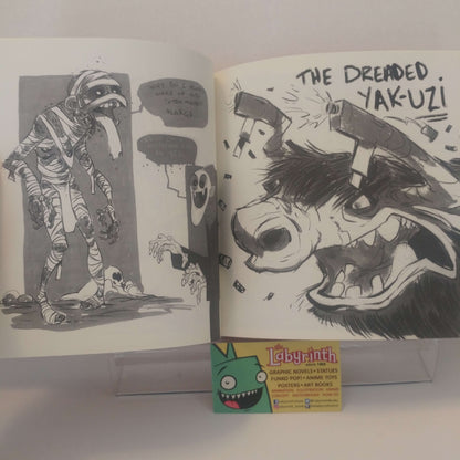 An open illustrated art book or comic spread on a white surface, showing black-and-white comic panels and a bold black ink illustration of a dog with the text 'THE DREMEID. YAK-UZ!'.