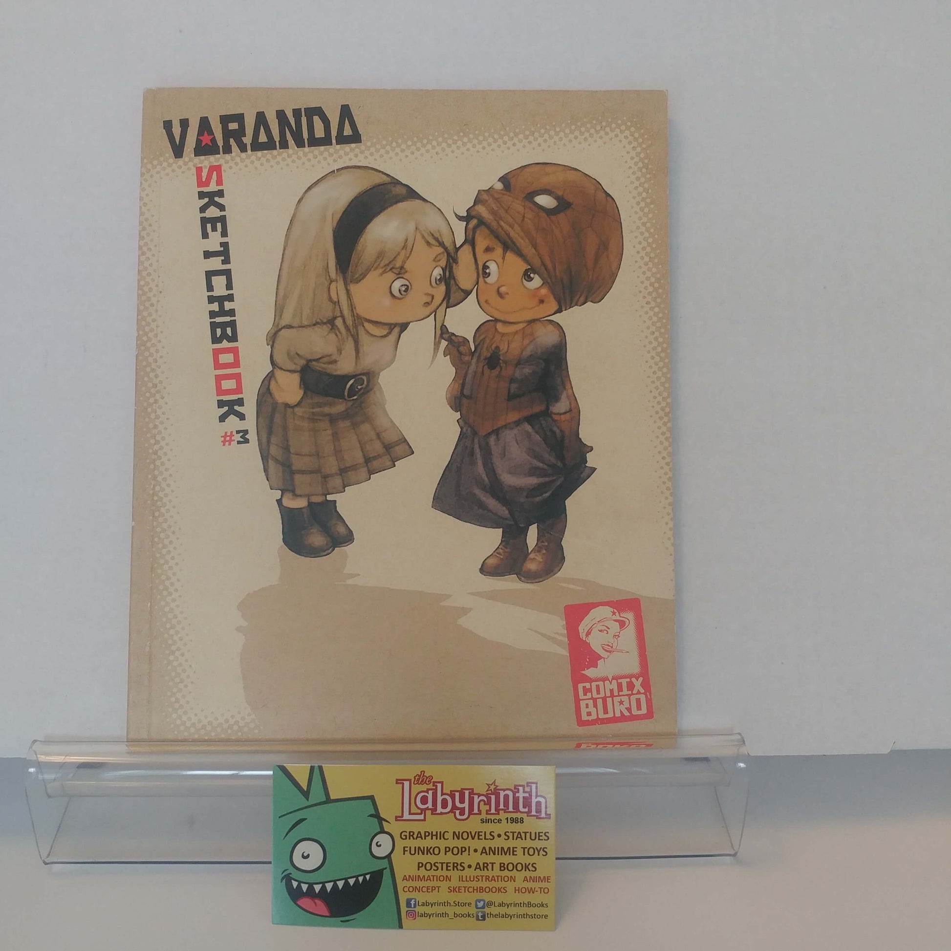 A comic book or sketchbook titled 'VARANDA SKETCHBOOK' featuring two illustrated children, displayed on a clear stand with a Labyrinth business card in front against a plain light background.
