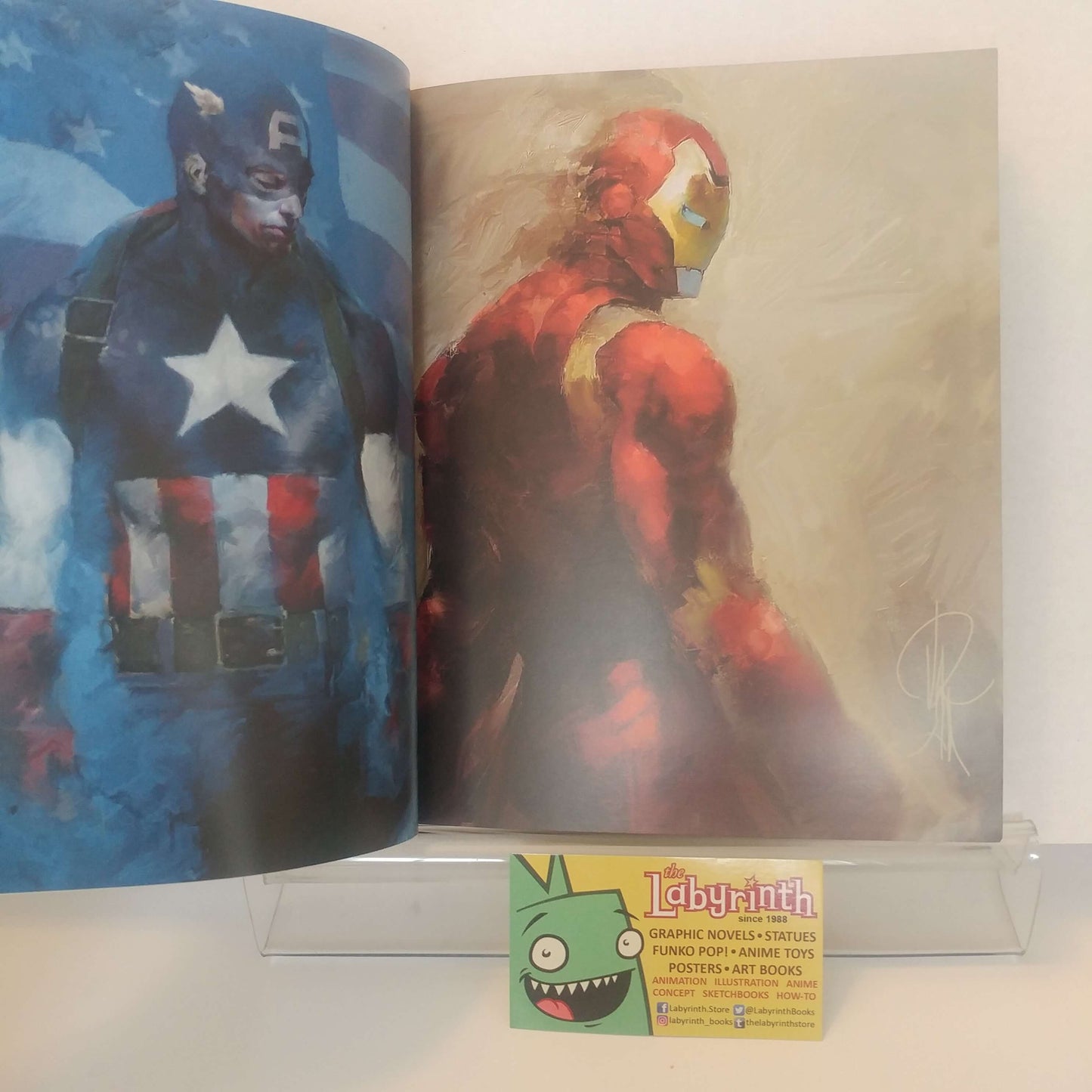 An open art book or portfolio displayed on a clear acrylic stand, showing two illustrated pages with Marvel characters: Captain America on the left and Iron Man on the right. A small card with the Labyrinth logo is placed in front of the book.