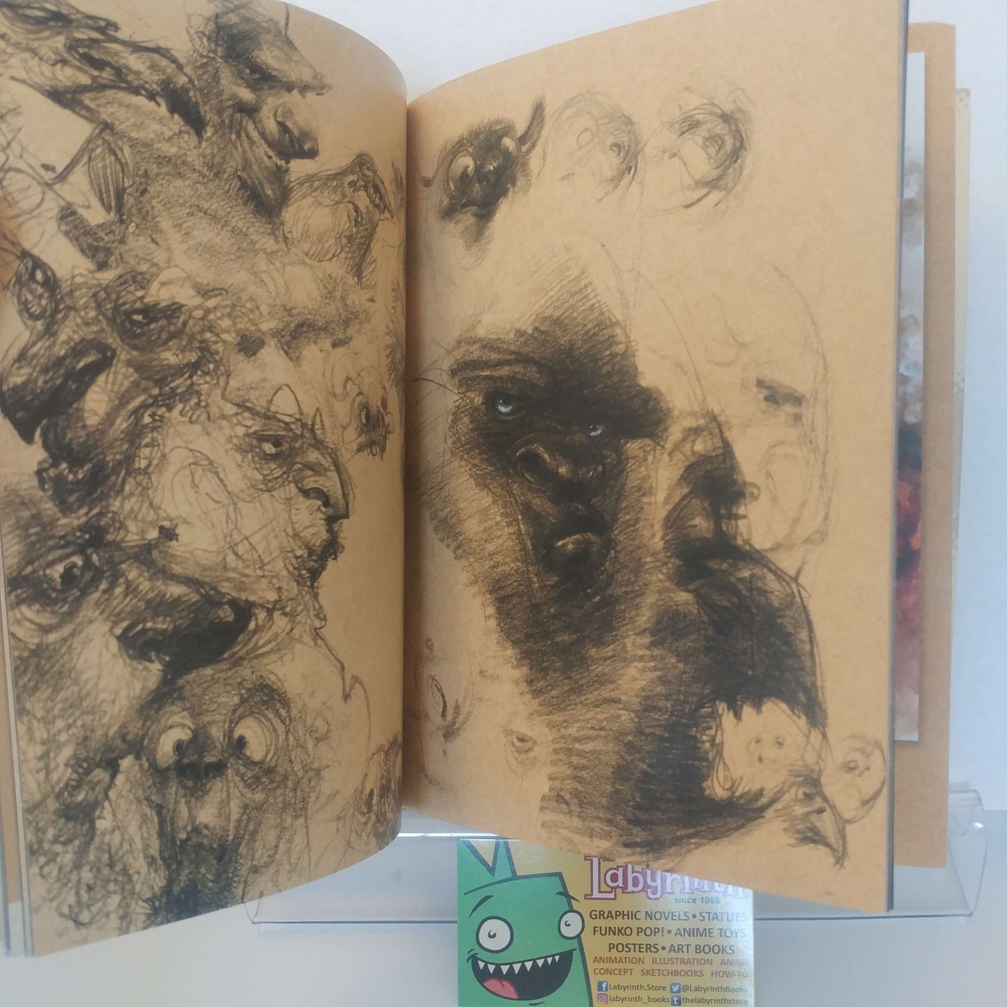 An open sketchbook displaying detailed black ink and charcoal drawings of faces on aged beige paper, with a small green cartoon card visible at the bottom.