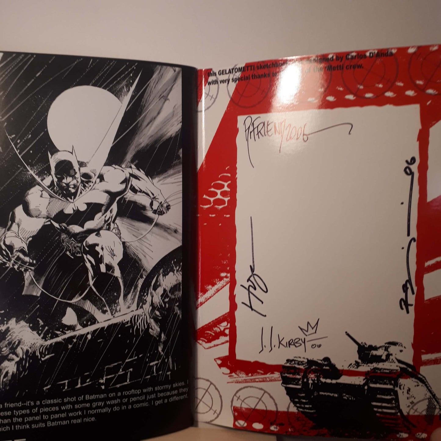 An open book or printed booklet showing a black-and-white comic-style Batman illustration on the left page and a red-bordered autograph page on the right, with signatures and a tank illustration at the bottom.
