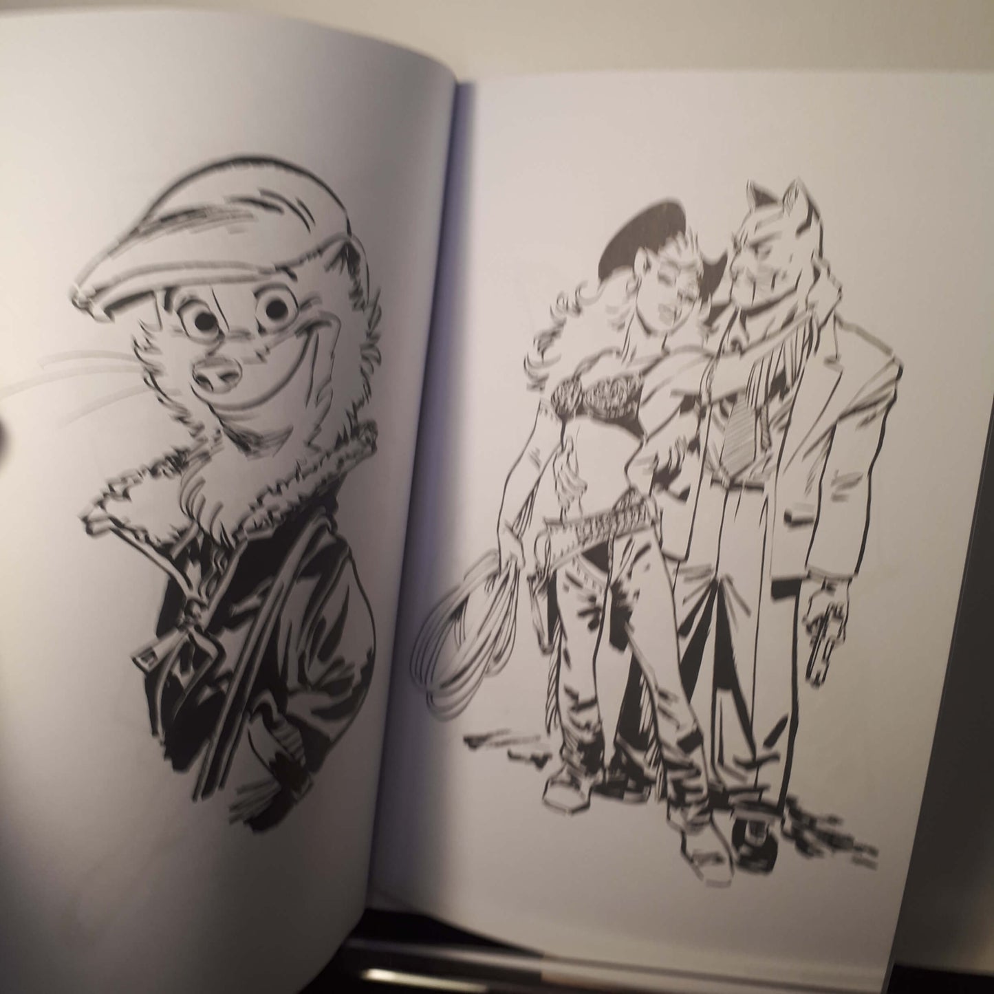 An open sketchbook or art book showing black ink illustrations on white pages. The left page has a cartoon animal portrait wearing a cap, and the right page shows a group of illustrated characters in a dynamic scene.