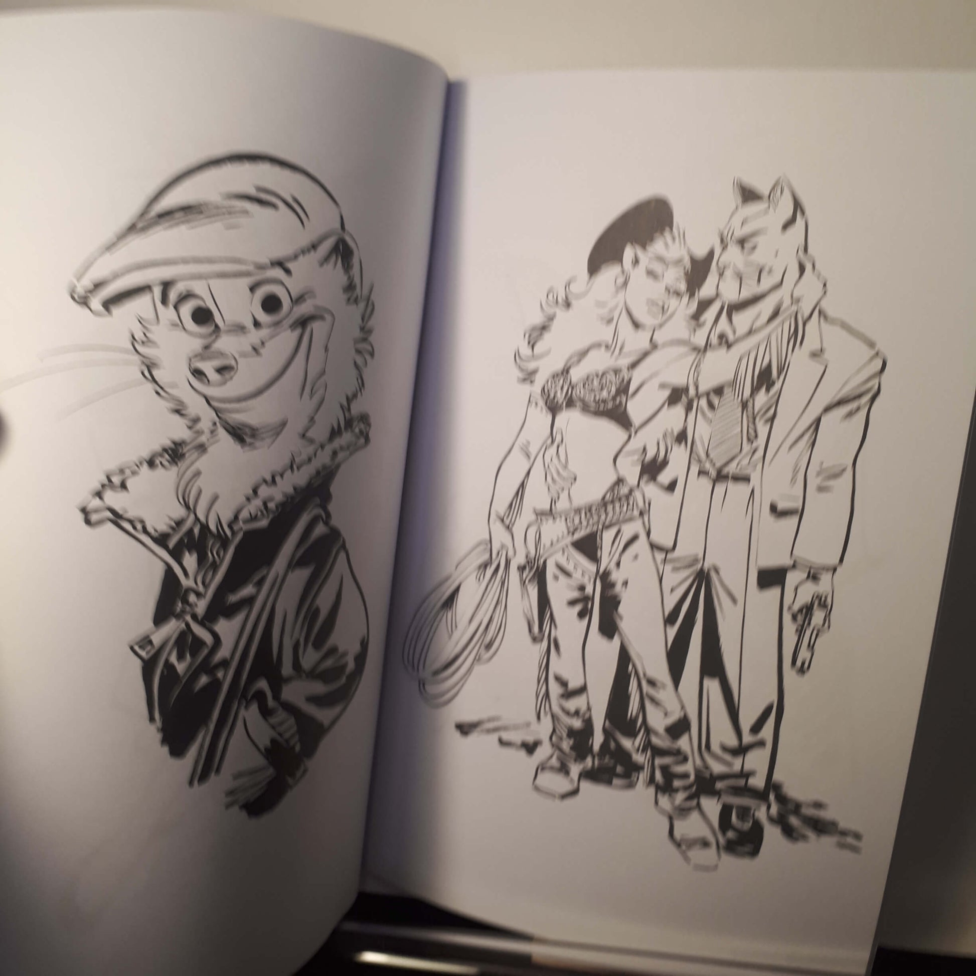 An open sketchbook or art book showing black ink illustrations on white pages. The left page has a cartoon animal portrait wearing a cap, and the right page shows a group of illustrated characters in a dynamic scene.