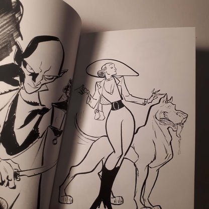 A close-up of an open sketchbook or art book showing black ink line drawings on white paper. The right page depicts a woman in a wide-brimmed hat and coat standing beside a large dog, while the left page shows part of another illustrated figure.