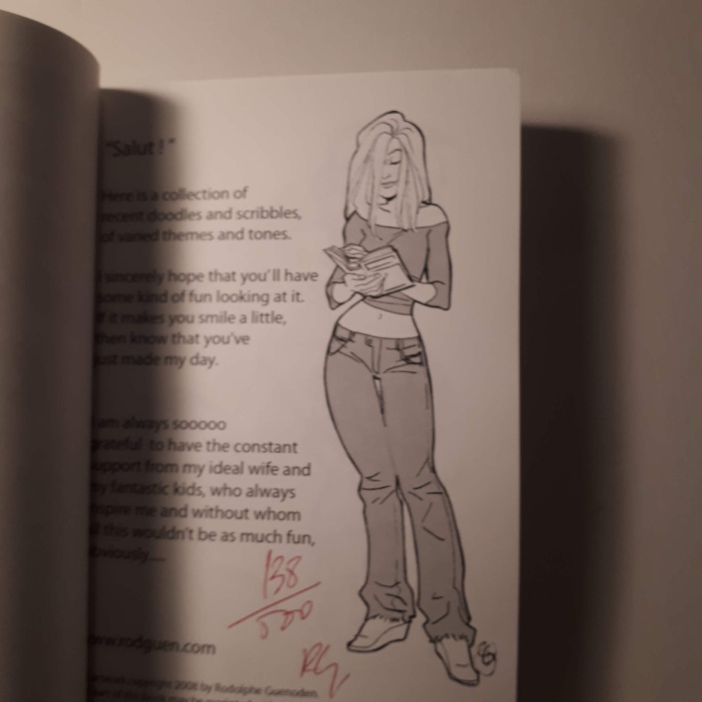 A printed page from a book or comic featuring a black-and-white line drawing of a woman holding a book, with printed text on the left and a red handwritten signature near the bottom.