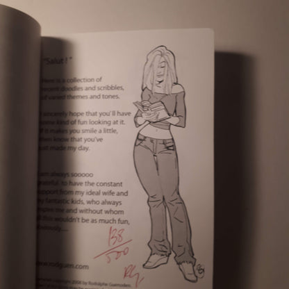 A printed page from a book or comic featuring a black-and-white line drawing of a woman holding a book, with printed text on the left and a red handwritten signature near the bottom.