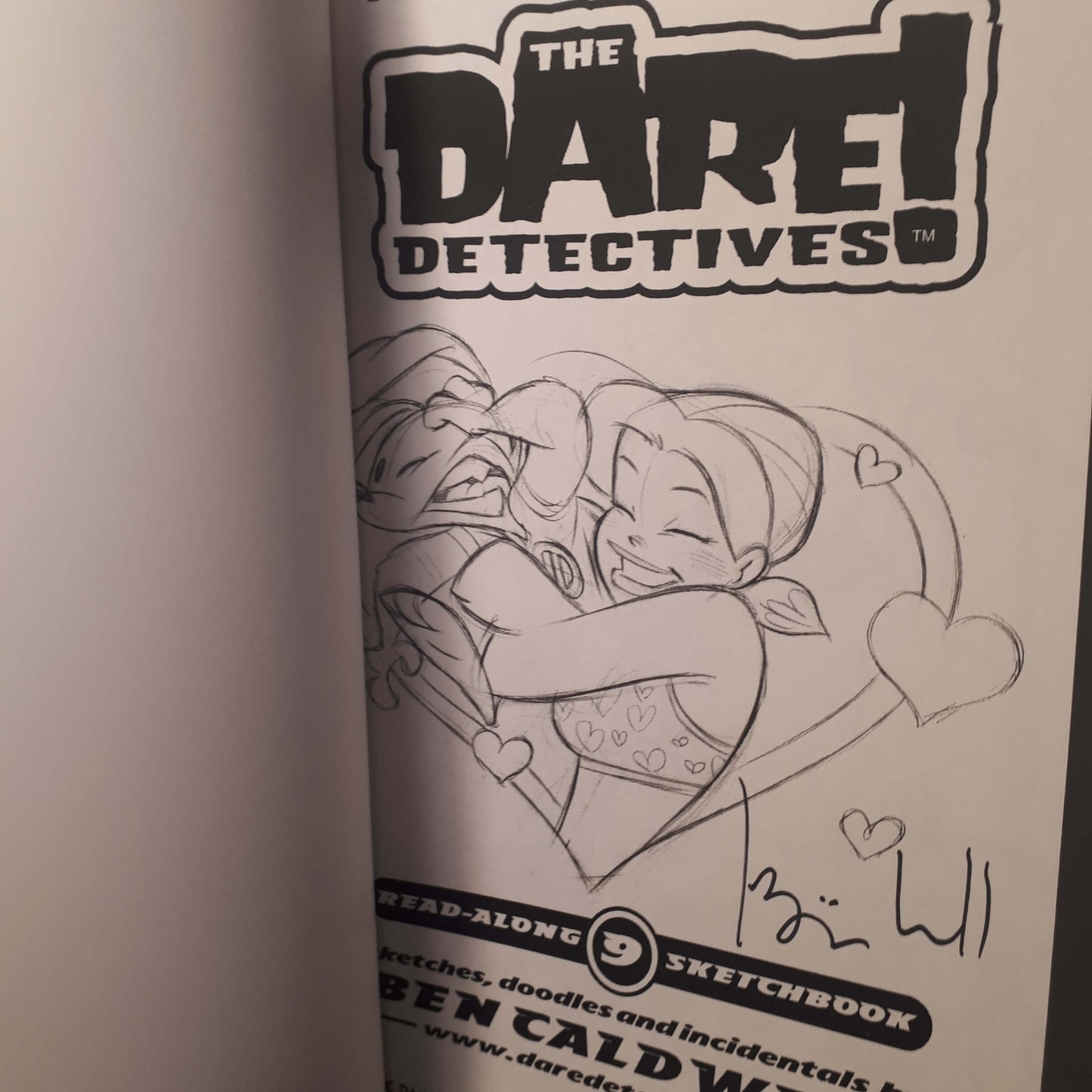 A close-up of a printed sketchbook page from 'The Dare Devoted' featuring a black-and-white line drawing of two characters hugging, with the title logo at the top and a handwritten signature near the bottom.