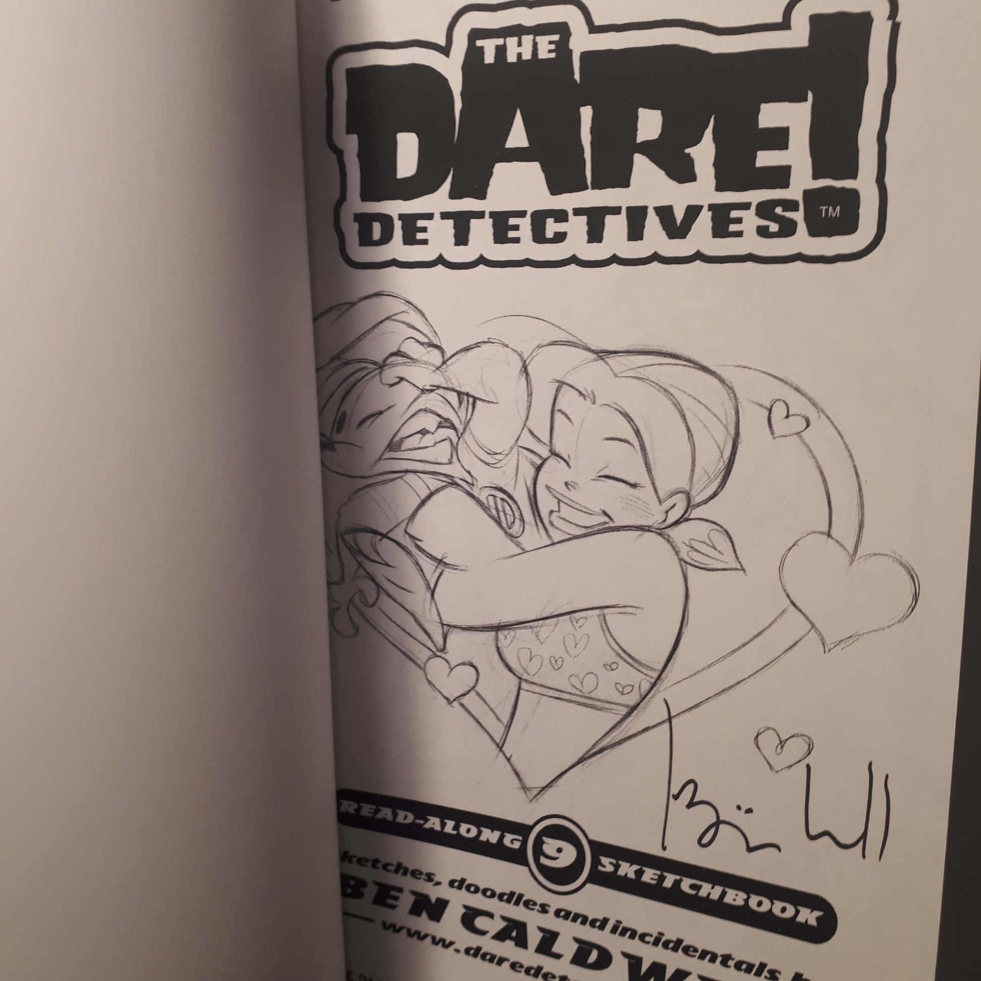 A close-up of a printed sketchbook page from 'The Dare Devoted' featuring a black-and-white line drawing of two characters hugging, with the title logo at the top and a handwritten signature near the bottom.