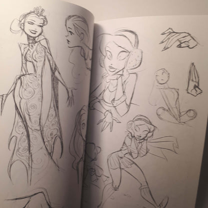 An open sketchbook showing pencil character designs and costume drawings on white pages, photographed indoors with a warm light source.