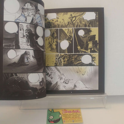 An open comic book or graphic novel displayed upright on a clear acrylic stand against a white wall. The pages show black-and-white and yellow-toned illustrated panels with speech-bubble circles cut out, and a small card labeled 'Labyrinth' is visible below the stand.