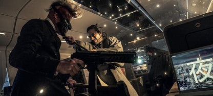 A dramatic cinematic still from a film showing two men in suits holding a large gun in a dark, high-tech interior with ceiling lights and a screen displaying Japanese characters.