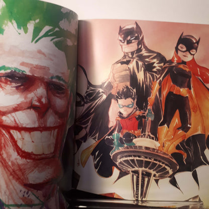 An open art book or magazine showing printed illustrations, including a large Joker face on the left page and Batman, Robin, and Batgirl characters on the right page.