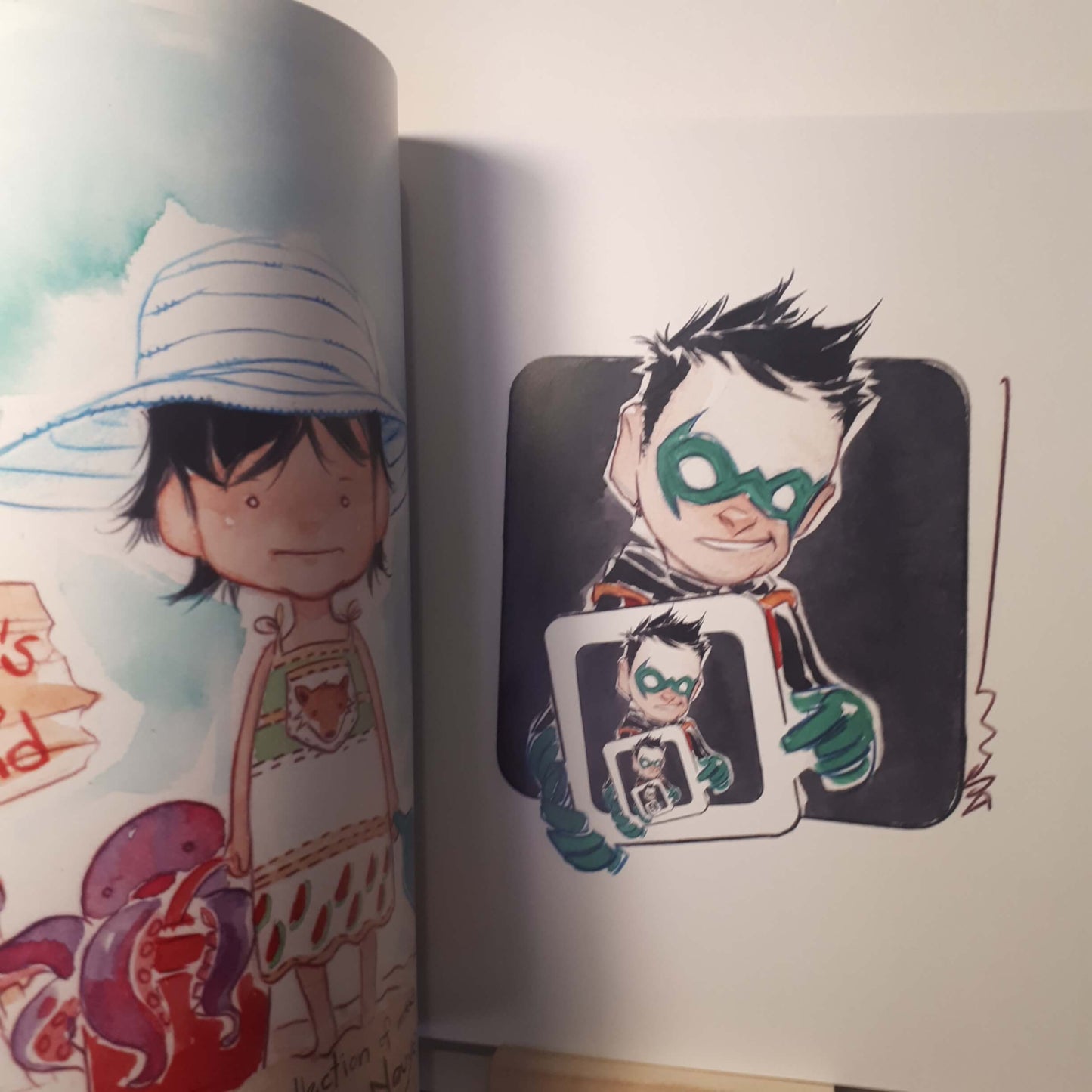 An open art book or sketchbook showing illustrated pages. The left page has a watercolor-style drawing of a person in a striped hat, and the right page shows a comic-style character with a green mask holding a mirror-like frame with a smaller version of the same character.