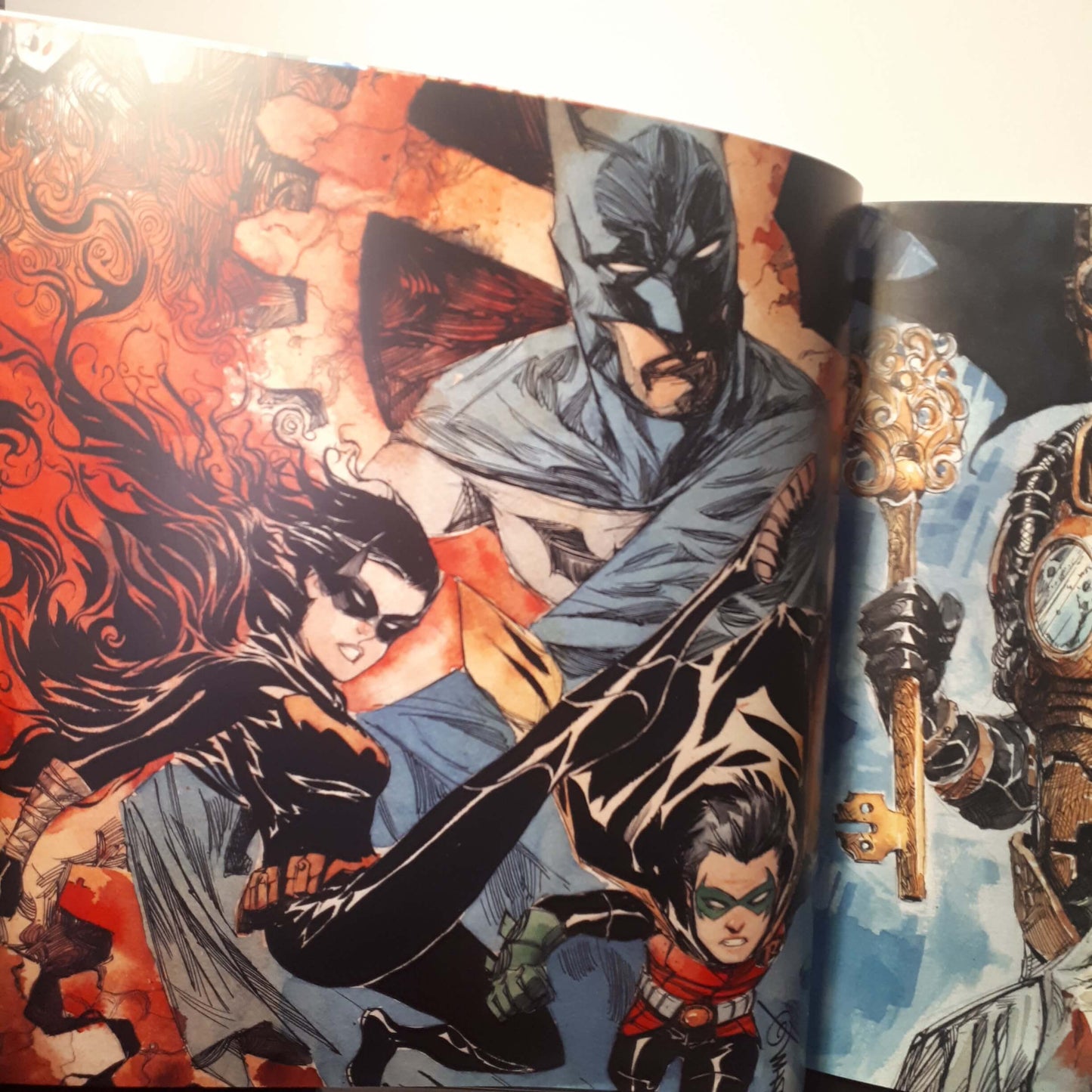 An open comic book or art book showing a dynamic illustrated cover with Batman, Robin, and another character in action, with a partially visible right page featuring a steampunk-style figure.