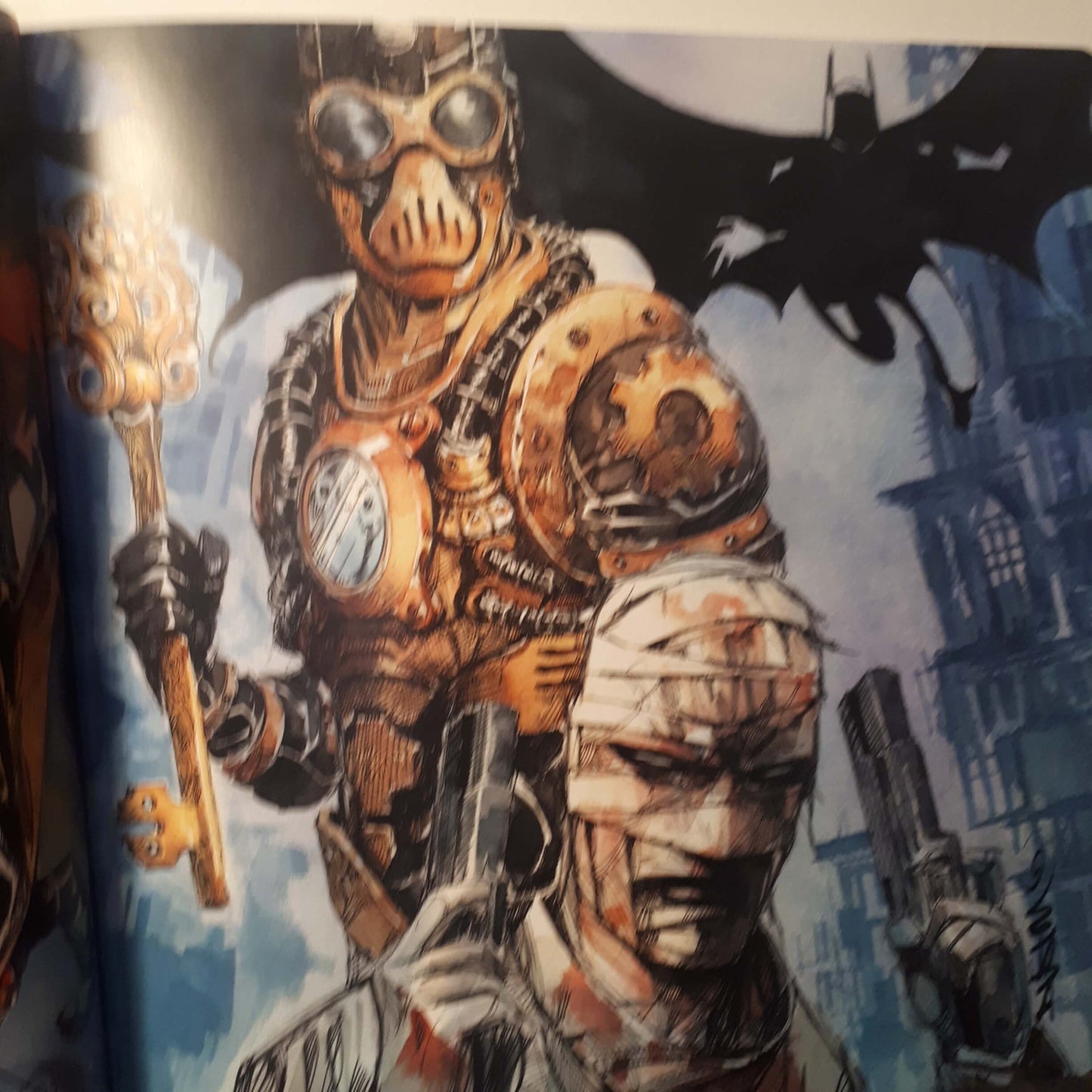 A close-up of printed artwork showing a steampunk-style armored figure with goggles and a key, alongside a bandaged mummy-like character, against a blue-gray city background with a bat silhouette.
