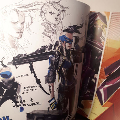 A printed art book or concept art portfolio page showing character sketches and annotations, with one illustrated figure in blue and black armor holding a large weapon. The page is photographed at an angle with other printed pages visible on the right.
