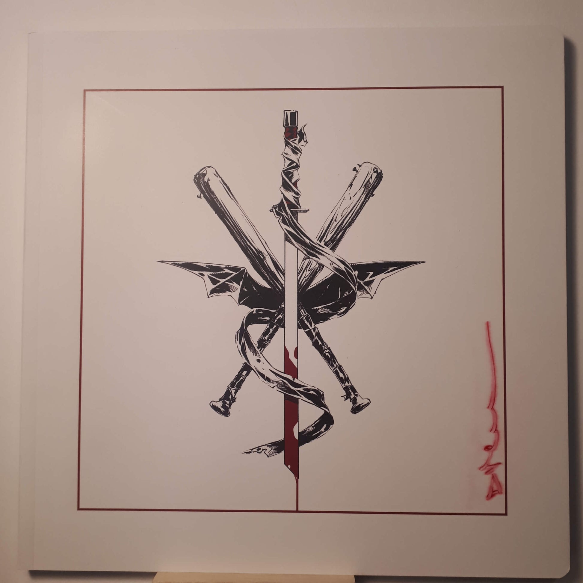 A square art print or poster featuring a black ink illustration of crossed swords and a snake wrapped around a vertical blade, set within a thin red border on a white background. A red signature or mark appears on the lower right side.