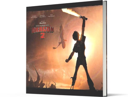 A hardcover book or album cover for 'How to Train Your Dragon 2' shown at an angle against a plain white background. The cover features a dramatic silhouette of a dragon rider holding a flaming sword against a bright orange and yellow sky, with the title 'How to Train Your Dragon 2' and the 'DreamWorks' logo visible.