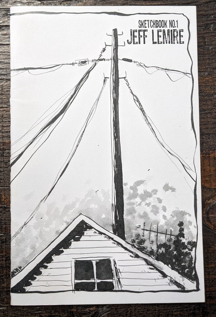 A sketchbook featuring a black and white hand-drawn illustration of a house roof, power lines, and a utility pole, with the text 'SKETCHBOOK NO.1 JEFF LEMIRE' in the top right corner, placed on a dark wooden surface.